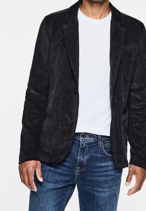 Hombre con blazer de pana negro, camiseta blanca de cuello redondo y vaqueros azules desgastados, de pie con los brazos relajados.
