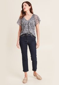Vloeiende zwart-witte bloemenblouse met korte, doorschijnende mouwen, gecombineerd met donkerblauwe, tapered jeans en lichtgouden instap-schoenen.
