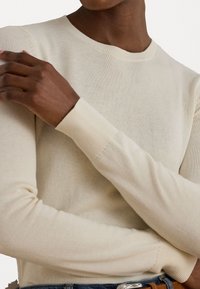 Beige strikket langermet topp med rund halslinning. Myk tekstur, tettsittende fasong og ribbede mansjetter. Brukt sammen med jeans og et belte.
