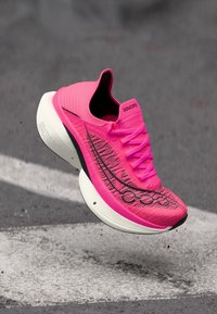 Φωτεινό ρόζ αθλητικό παπούτσι Saucony με λευκή παχιά σόλα, μαύρες λεπτομέρειες και κορδόνια, αιωρούμενο πάνω από επιφάνεια ασφάλτου.