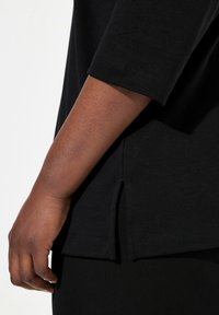 Top nero a maniche corte con dettaglio laterale a spacco. Realizzato in tessuto morbido, presenta un orlo arrotondato e maniche a cappotto per una vestibilità comoda.