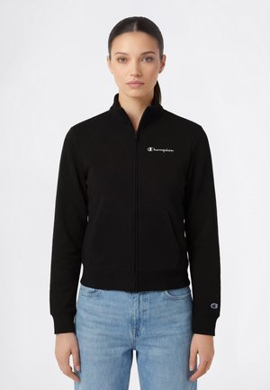 Femme debout, face à l'avant, portant une veste noire zippée avec des logos Champion sur la poitrine et la manche, associée à un jean bleu clair.