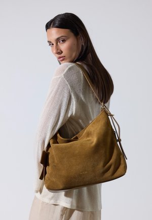 Žena s dugom tamnom kosom koja nosi kremasti pleteni džemper i nosi veliku smeđu torbu od brušene kože preko desnog ramena.