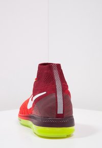 Červená sportovní bota s pleteným designem, vysokým nákolením a průhlednou zelenou podešví. Má bílé logo Nike a texturované detaily.