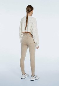 Mujer con cabello largo trenzado, vistiendo un suéter corto de punto blanco, jeans ajustados beige y zapatillas blancas, de pie con la espalda hacia el espectador.
