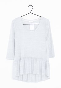 Haut à manches longues gris clair avec un décolleté arrondi, un ourlet volanté et une texture de tissu douce. Accroché à un crochet noir contre un fond blanc.