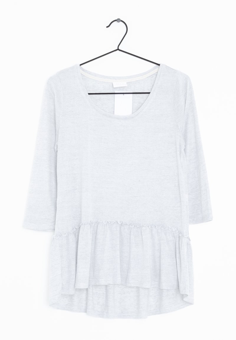 Haut à manches longues gris clair avec un décolleté arrondi, un ourlet volanté et une texture de tissu douce. Accroché à un crochet noir contre un fond blanc.