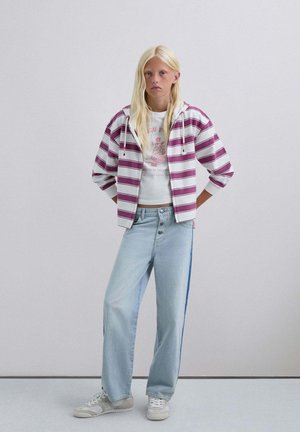 Sudadera corta de rayas moradas y blancas sobre una camiseta gráfica blanca, combinada con vaqueros de pierna ancha azul claro con una franja lateral azul y zapatillas blancas.