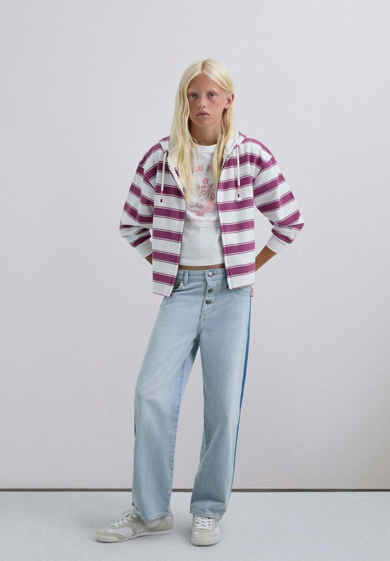 Sudadera corta de rayas moradas y blancas sobre una camiseta gráfica blanca, combinada con vaqueros de pierna ancha azul claro con una franja lateral azul y zapatillas blancas.