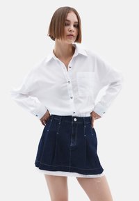 Witte langmouwige overhemd met een borstzak, gecombineerd met een donkere denim geplooide rok met een ruwe zoom en een knoopsluiting aan de voorkant.