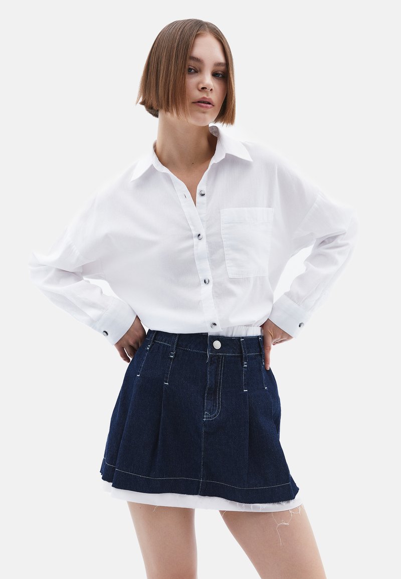 Witte langmouwige overhemd met een borstzak, gecombineerd met een donkere denim geplooide rok met een ruwe zoom en een knoopsluiting aan de voorkant.