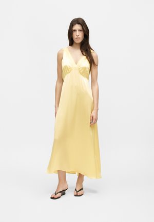 Femme portant une robe midi en satin jaune pâle sans manches avec un col en V et des sandales noires à talons sur un fond clair uni.