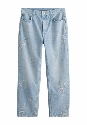 Hellblaue Jeans mit weißer Blumenstickerei, getragen mit einer weißen, strukturierten Langarmbluse vor einem schlichten Hintergrund.