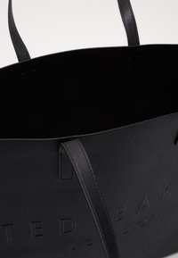 Czarna torba typu tote z fakturowanej skóry, z cienkimi uchwytami i wytłoczonym logo "TED BAKER LONDON" z przodu na jednolitym tle.