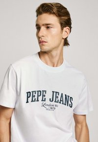 Baltos medvilninės marškinėliai su tamsiai mėlyna "PEPE JEANS London W11 1973 Portobello Rd" užrašu. Apvalus kaklas ir trumpomis rankovėmis.