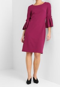 Lila klänning med rund hals och klockärmar. Slät, texturerad tyg, knälång design, kombinerad med svarta högklackade skor.