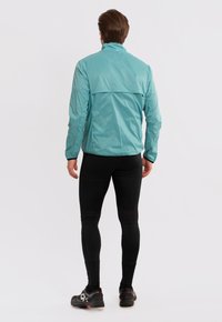Lichte, turquoise jas met een hoge kraag, voorzien van een rugventilatie detail, gedragen over zwarte leggings en schoenen. Glad, waterafstotend materiaal.