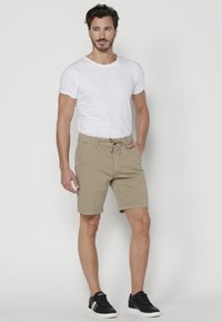 Vīrietis baltā T-kreklā, bēšās biksēs ar aukliņu un melnās sporta kurpēs stāv ar vienu roku kabatā pret vienkāršu fona.