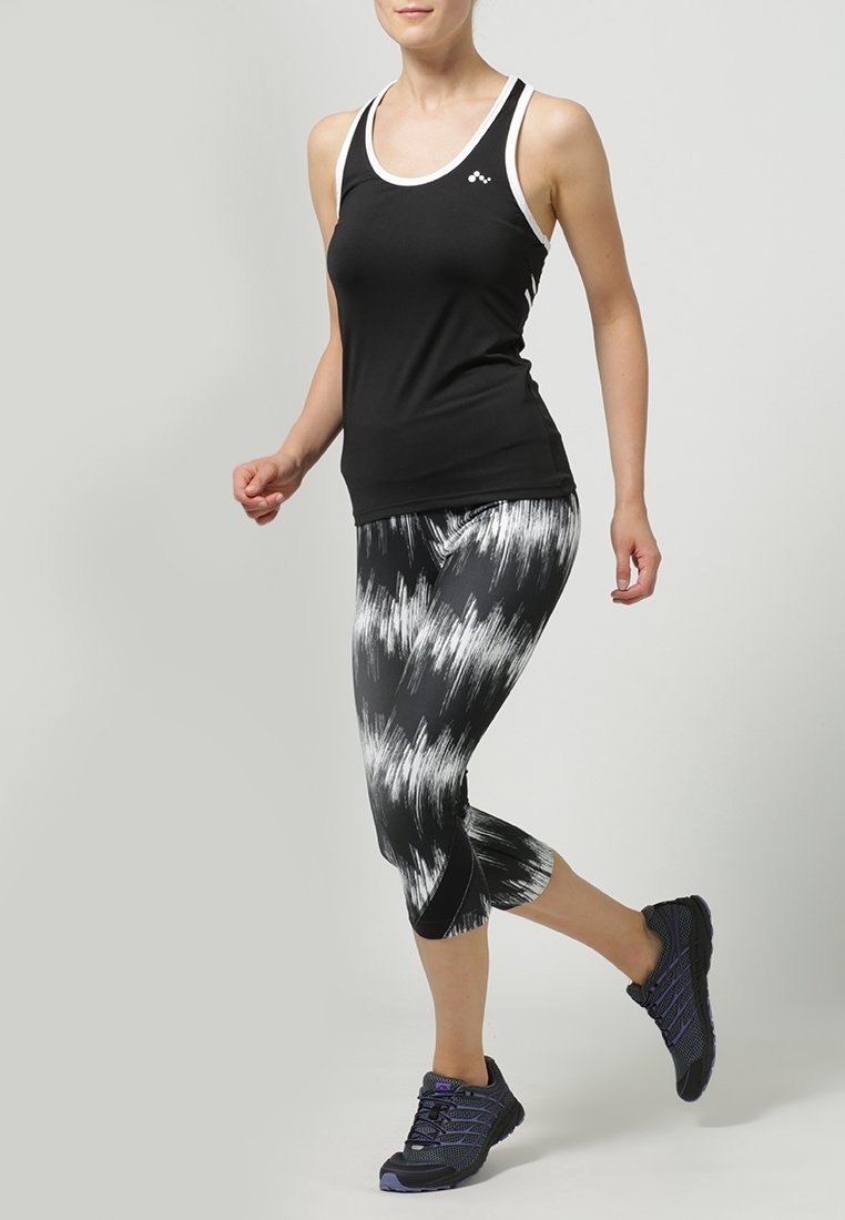 Czarny sportowy top na ramiączkach z białymi wykończeniami, zestawiony z legginsami 3/4 w czarno-białe wzory oraz czarnymi butami sportowymi.