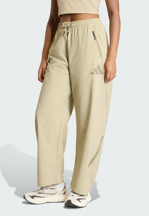 Beige sportbroek met een elastische tailleband, trekkoord, zijzakken en subtiele branding op de dij, gedragen met witte sneakers.