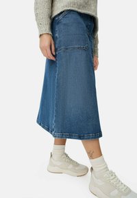 Jupe midi en denim dans un lavage bleu foncé, avec une coupe droite, des poches latérales et un ourlet brut. Portée avec des baskets beige clair et des chaussettes.