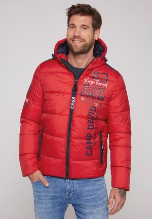 Lachende man met een rood gewatteerd jack met tekst, blauwe rits, capuchon en lichtblauwe jeans, staand met één hand in de zak.