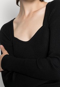 Svart ribbat stickat linne med fyrkantig halsringning, långa ärmar och texturerad mönster. Tyget är stretchigt, vilket skapar en figurnära siluett.