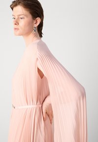 Light pink pleated dress with flowing sleeves, a fitted waist belt, and a soft texture. Complemented by round metallic earrings.