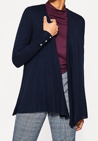 Marineblauer langer Cardigan aus leichtem Strickstoff, mit drapiertem Vorderteil, Knopfdetails an den Ärmeln, kombiniert mit einem burgunderfarbenen Rollkragenpullover und karierten Hosen.
