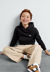 Black hooded sweatshirt, beige wide-leg pants, and brown and white sneakers. The child is seated with a playful expression.