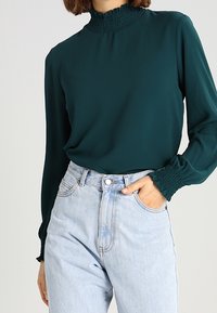 Blusa lunga teal con colletto smock alto, abbinata a jeans azzurri a vita alta con chiusura a bottone classica e tasche.