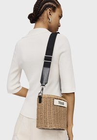 Borsa a tracolla in paglia intrecciata con una tracolla regolabile crema e nera, caratterizzata da un'etichetta bianca con "TOUS" in grassetto di colore nero.