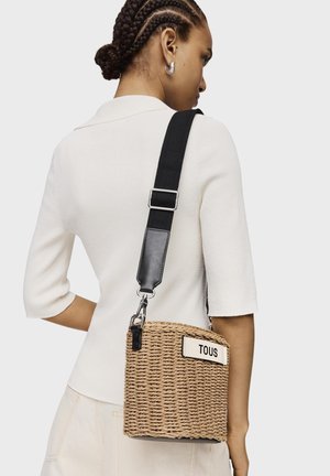 Sac bandoulière en paille tissée avec une sangle réglable crème et noire, arborant une étiquette blanche avec 'TOUS' en lettres noires en gras.