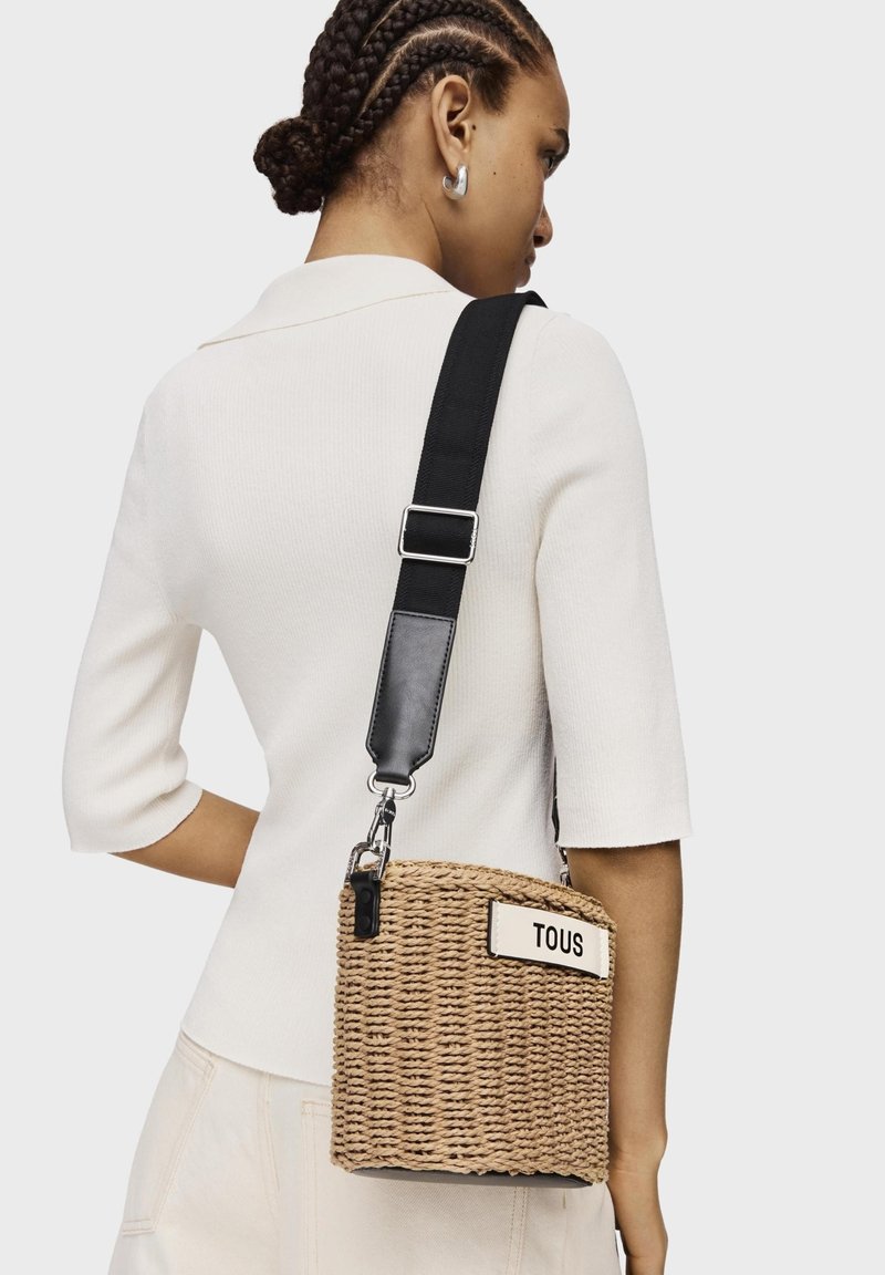 Borsa a tracolla in paglia intrecciata con una tracolla regolabile crema e nera, caratterizzata da un'etichetta bianca con "TOUS" in grassetto di colore nero.