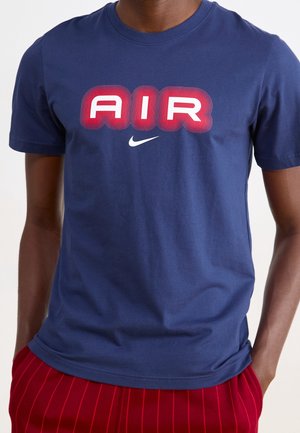 Man draagt een marineblauw t-shirt met rode en witte tekst "AIR" en een witte Nike swoosh-logo, gecombineerd met rode broeken met dunne verticale strepen.
