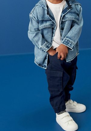 Barn iført en lyseblå denimjakke, hvid t-shirt, marineblå cargobukser og hvide velcro-sneakers, stående mod en ensfarvet blå baggrund.