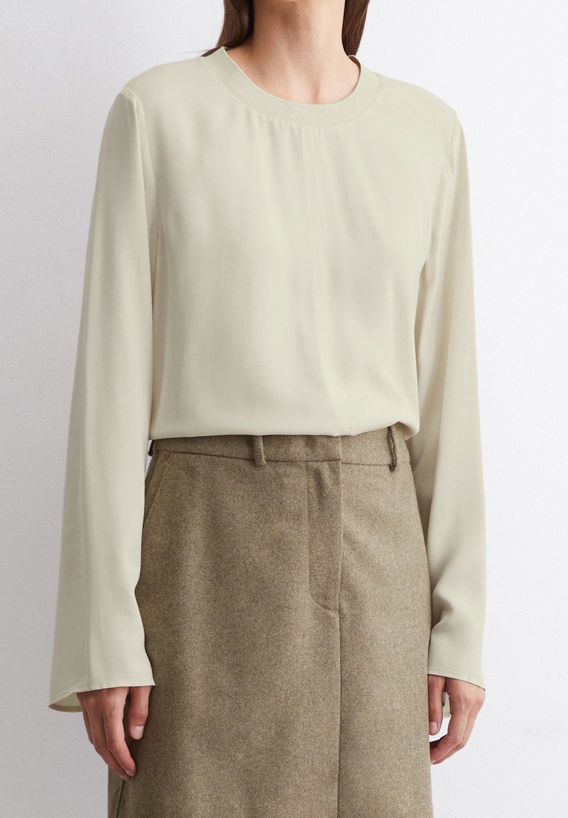 Helles beige Langarmbluse mit rundem Ausschnitt, glatter Textur und subtilen Faltendetails, kombiniert mit hochtaillierten braunen, maßgeschneiderten Hosen.