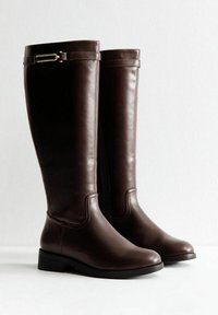 Tall dark brown leather boots with low heels and decorative straps near the top, standing side by side on a white surface.