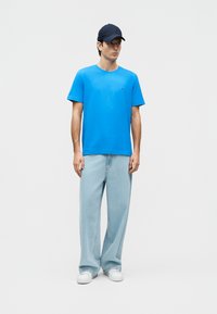 Νεαρός άνδρας που φοράει ένα φωτεινό μπλε T-shirt, ανοιχτό μπλε φαρδιά τζιν, λευκά αθλητικά παπούτσια και μαύρο καπέλο μπέιζμπολ, στέκεται με χαλαρά τα χέρια.