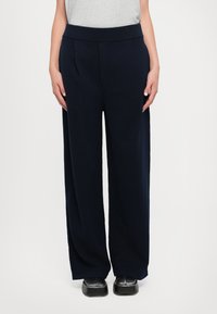 Pantalones de pierna ancha en tono marino con una cinturilla acanalada y pliegues frontales, confeccionados en una tela suave. Combinados con calzado negro plano.