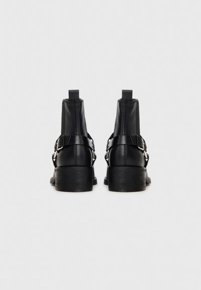 Par de botas de cuero negras hasta el tobillo con correas de hebillas plateadas, tacones cuadrados y lengüetas para tirar, vistas desde la parte trasera sobre un fondo blanco liso.