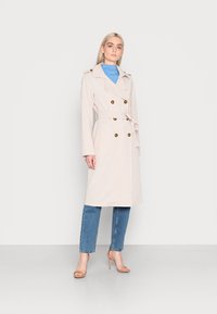 Beige, doppelt geknöpfter Trenchcoat mit einem Bindegürtel, ausgestattet mit goldfarbenen Knöpfen, Seitentaschen und einer lockeren Passform, getragen über einem blauen Oberteil und Jeans.