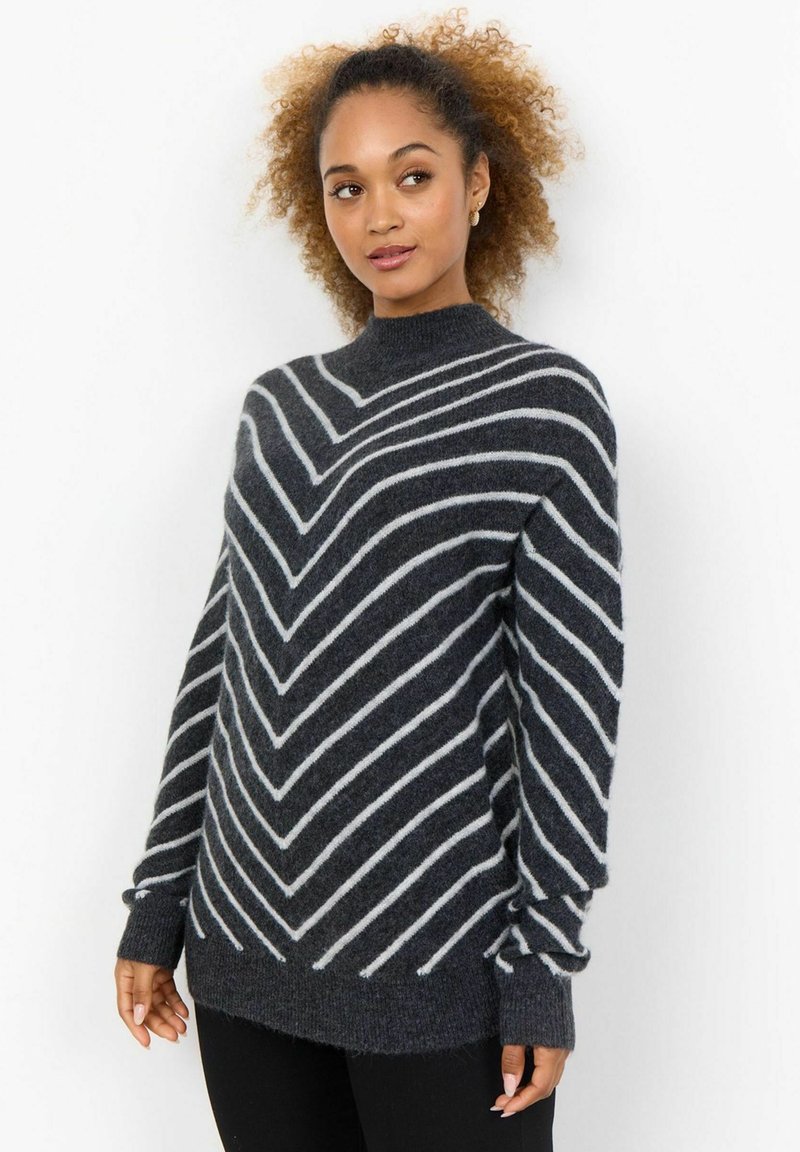 Pull en tricot gris foncé avec des rayures en chevron blanches, présentant un col montant et des manches longues. Texture douce et coupe décontractée.