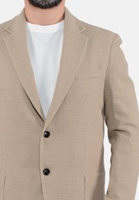 Blazer beige con texture, colletto a scollo, due bottoni neri e tasche laterali, indossato sopra una maglietta bianca a girocollo.