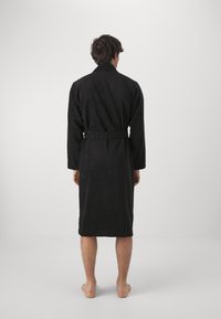 Peignoir noir texturé avec manches longues et ceinture, atteignant le mi-mollet, vu de dos, porté par une personne sans chaussures.