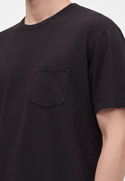Gros plan d'une personne portant un t-shirt noir uni à manches courtes avec une petite poche poitrine sur le côté gauche.