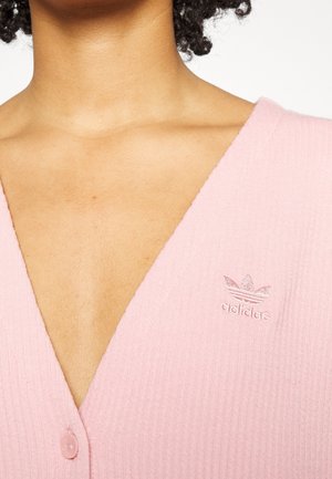 Zbliżenie osoby noszącej różowy prążkowany top z dekoltem w serek i małym haftowanym logo Adidas na piersi.