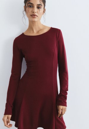 Femme aux cheveux foncés portant une robe moulante à manches longues de couleur rouge foncé, debout devant un fond clair uni.