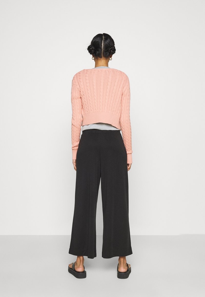Pull rose en maille torsadée, court, et pantalon large noir ample. Sandales à semelles épaisses. Texture lisse, design simple.
