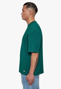 Grøn bomulds t-shirt med korte ærmer og rund halsudskæring. Har en lige pasform og en lille logo detalje nær kanten. Bæres med blå jeans.